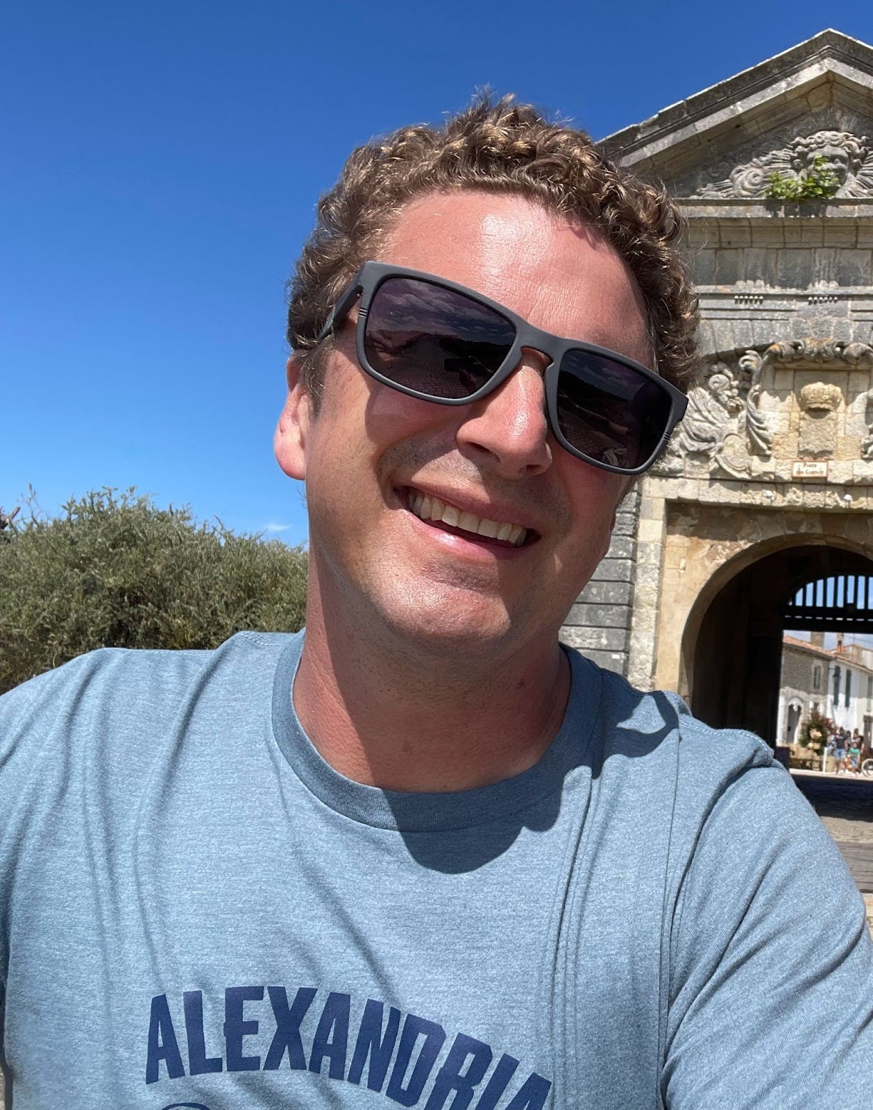 Head shot of white male with curly blond hair in sunglasses and a light blue t-shirt